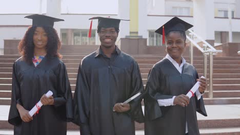 tres graduados universitarios afroamericanos están de pie afuera contra las escaleras. los estudiantes están vestidos con el uniforme festivo del maestro