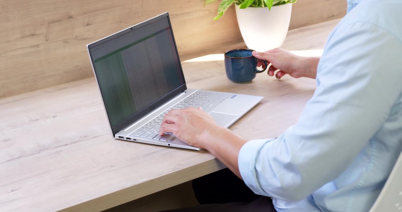 Asian man typing spreadsheet on laptop at desk, pausing work, sipping from blue mug, copy space