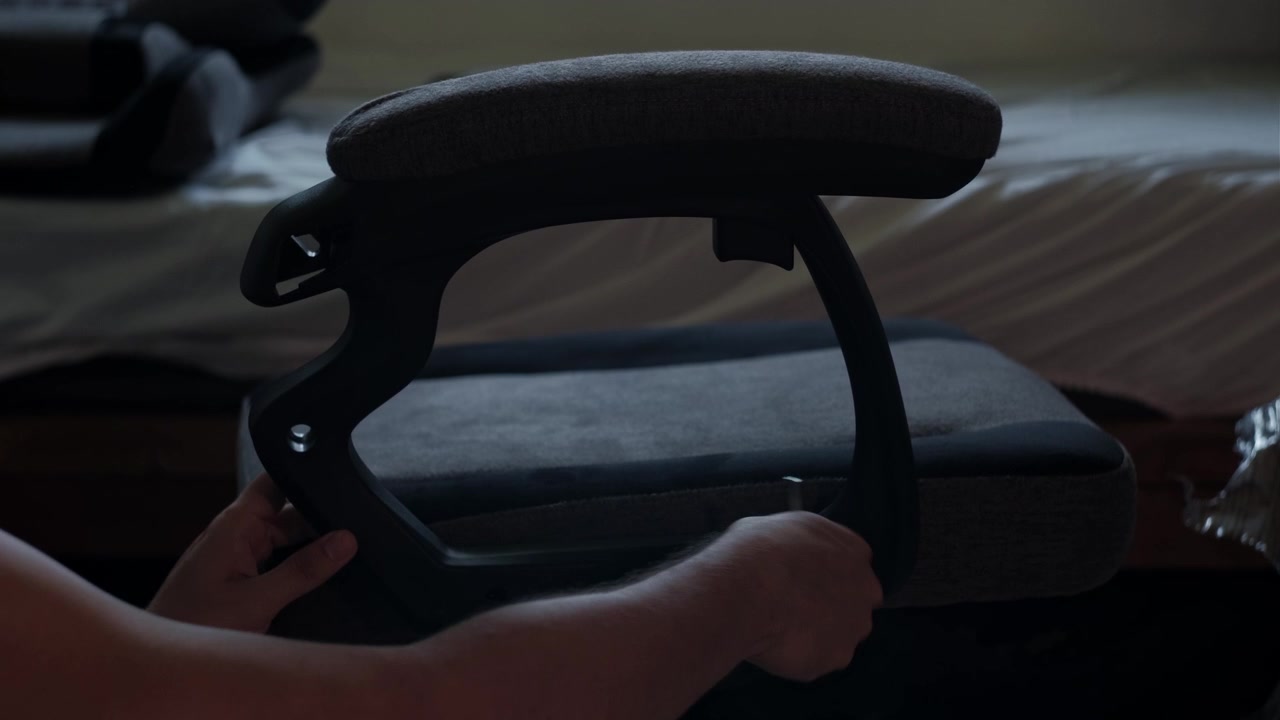 Inquadratura media di mani che allineano un telaio di bracciolo su una sedia da gaming in penombra durante l'assemblaggio di mobili da interno.