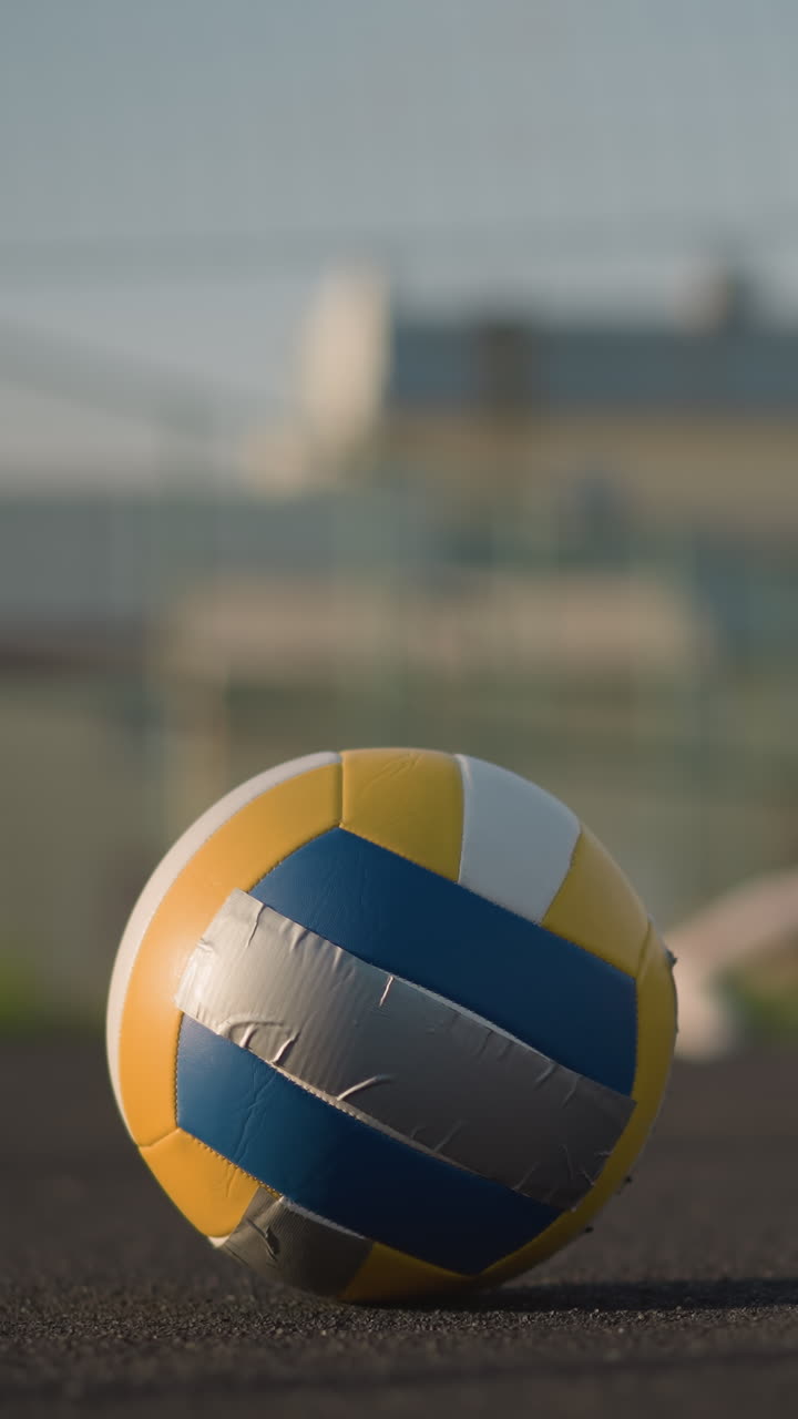 Primo piano di una palla da pallavolo a terra con sfondo sfocato che mostra atleti che si esercitano e fanno stretching all'aperto; sullo sfondo, un edificio è visibile con una vivace luce solare