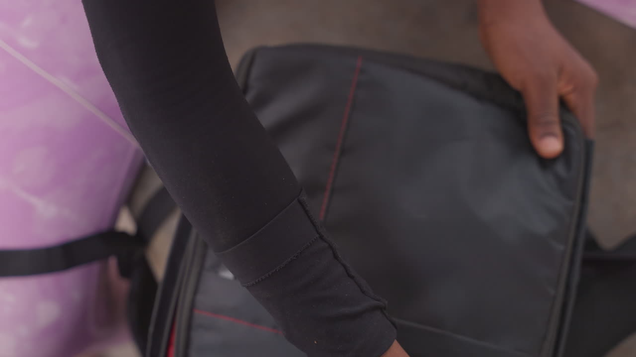 Primer plano de un fotógrafo negro empacando su mochila de cámara, manos acomodando lentes y tapas dentro del interior acolchado rojo, cerrando la mochila de nailon negro, con leggings morados visibles, una preparación concentrada para una sesión de fotos urbana.