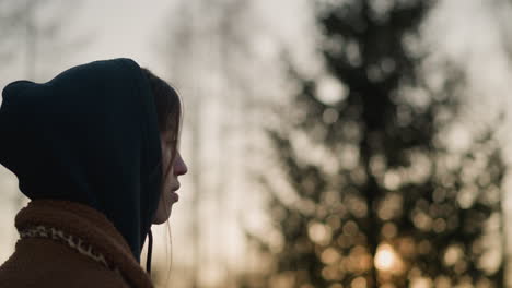 a melancholic girl walks through a park at sunset, wearing a brown coat and a hoodie with the hood up, partially concealing her face. blurred background of trees and fading sunlight