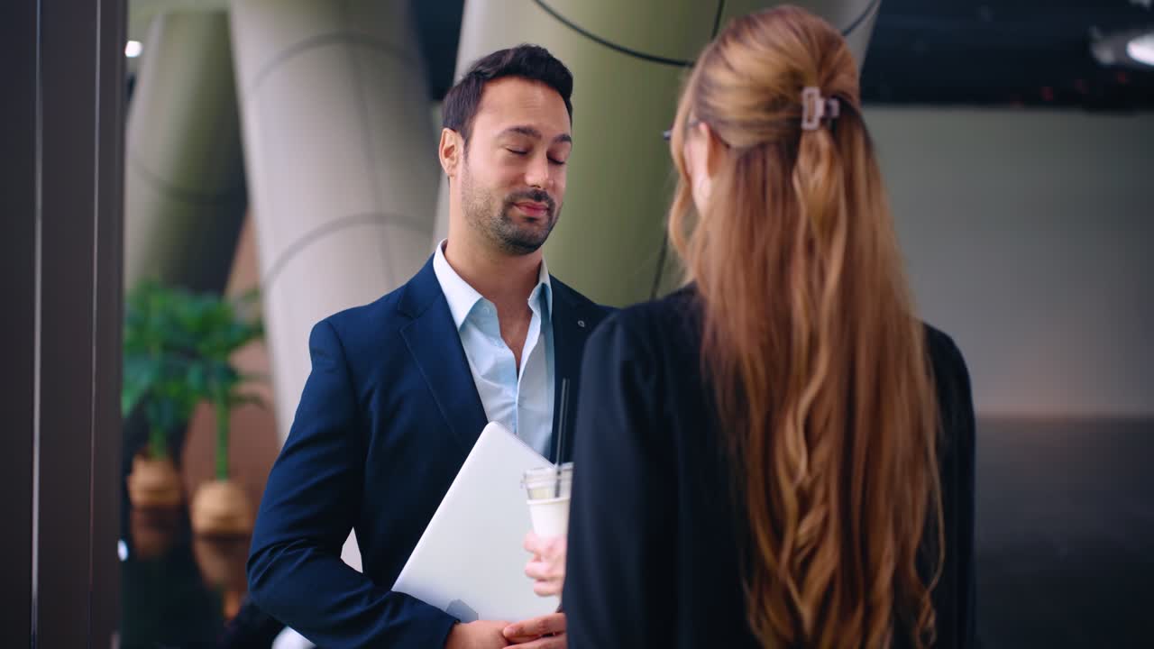 Two corporate colleagues standing in a modern office hall having a friendly small talk and discussing personal life during a work break while the man holds a laptop and smiles
