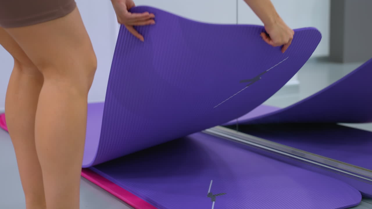 mujer atlética en ropa deportiva marrón recoge una alfombra de yoga púrpura en un estudio de fitness moderno, el espejo refleja sus acciones, creando una escena dinámica y atractiva de preparación para el entrenamiento