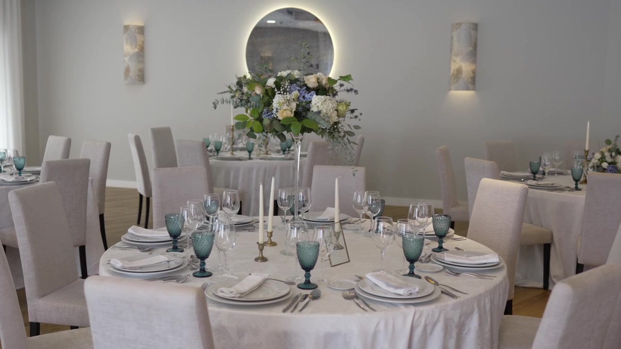 Elegante allestimento della tavola per un ricevimento di nozze con tovaglie bianche, alti centrotavola floreali di fiori bianchi e blu, e bicchieri, candele e stoviglie disposti con cura.