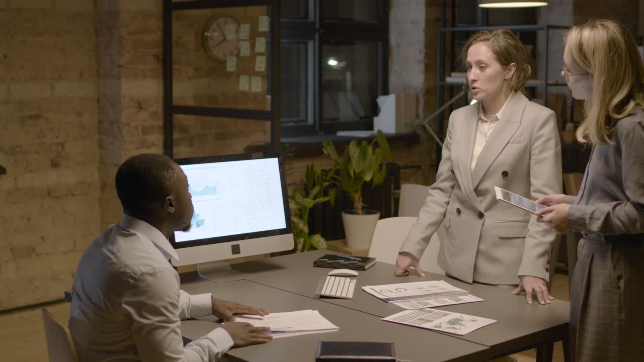 American Man Sitting At Desk In The Office While Talking About Graphics And Stadistics With Two Female Coworkers Who Are Standing