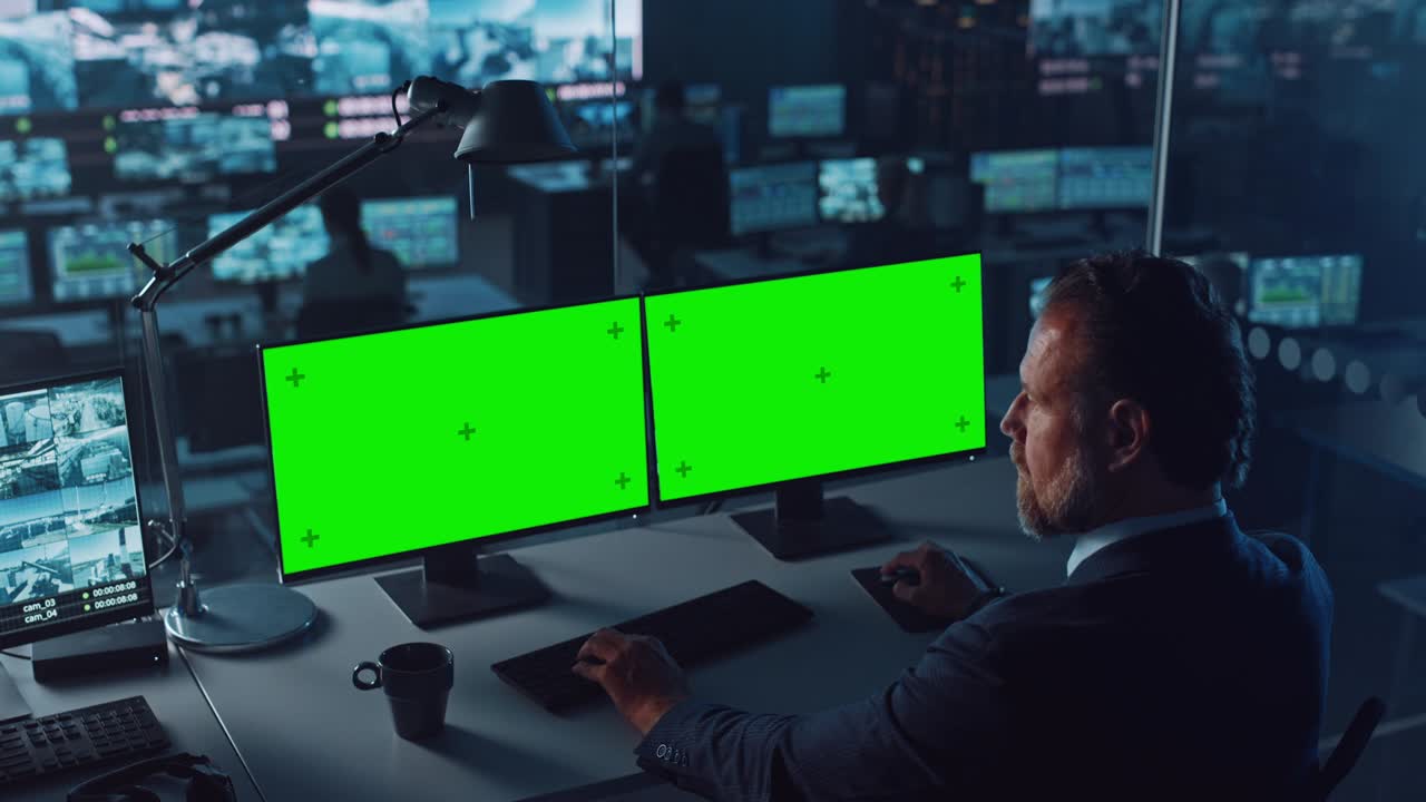 un oficial masculino trabaja en una computadora con dos maquetas de pantalla verde en un centro de vigilancia en un centro de monitoreo portuario con múltiples cámaras en una gran pantalla digital. los empleados se sientan frente a las pantallas.