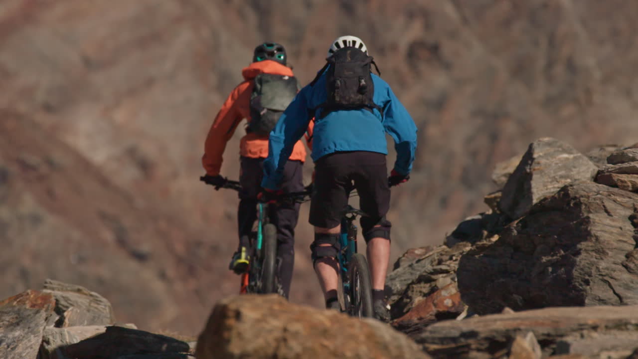 Dois ciclistas de montanha passando pela câmera e descendo a colina com amplas vistas do vale alpino em Saas-Fee, Alpes Suíços. Ação dinâmica na trilha, cenário de montanha pitoresco e clima cinematográfico de aventura de verão.
