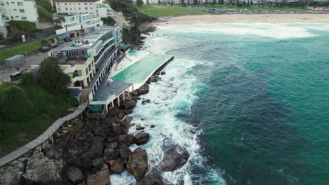 Aufschlussreiche-Drohnenaufnahme-Des-Bondi-Beach-Eisberg-Schwimmbads-Mit-Bondi-Beach-Im-Hintergrund-Bei-Sonnenuntergang