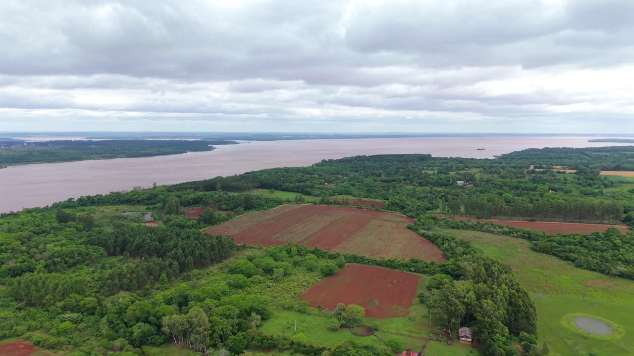A stunning aerial shot of bright red agricultural fields in Campichuelo, Paraguay, contrasting with green forests. The wide, brown Paraná River flows in the background