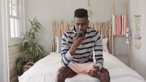 Happy-african-american-man-wearing-shorts-sitting-on-bed-talking-on-smartphone,-slow-motion