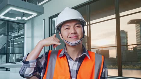 primer plano de ingeniero masculino asiático con casco de seguridad sonriendo a la cámara y haciendo el gesto de llamarme mientras está de pie con el proyecto de drones