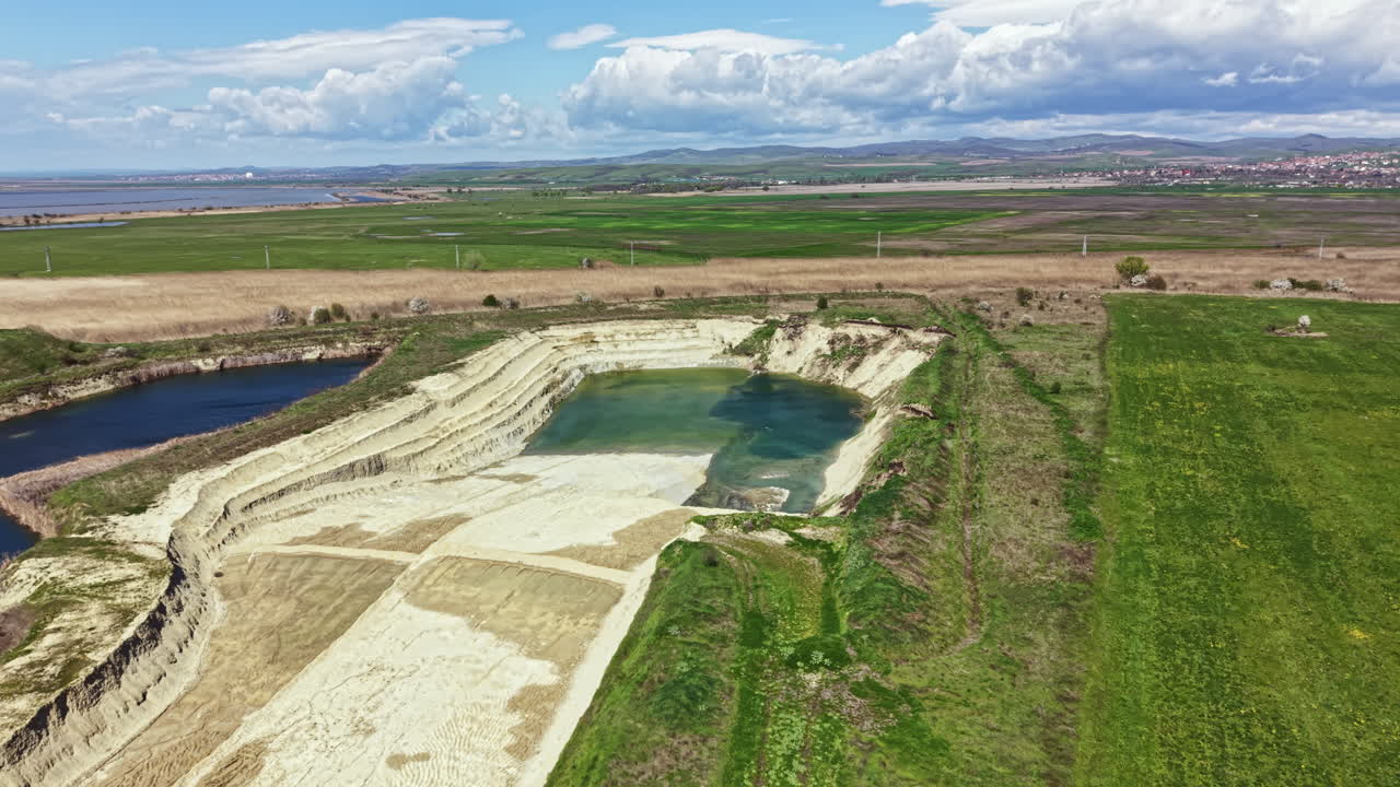 Drone view shows a large sand pit beside a river and green fields under a cloudy sky in a rural area with hills in the distance