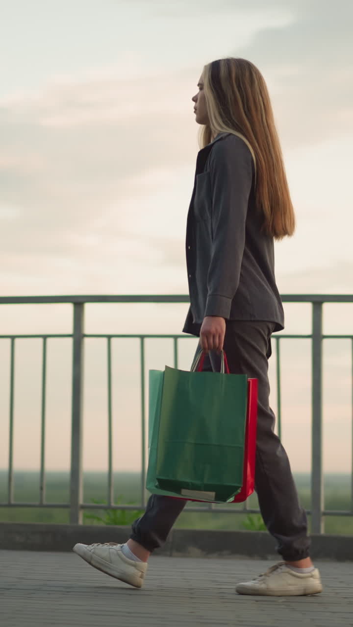 vista lateral de una dama con ropa gris caminando a lo largo de un carril de hierro con una bolsa de compras y una bolsa de mano negra por la noche, con luz natural suave y fondo borroso