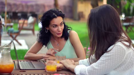 Two-women-sitting-at-the-outdoors-cafe---talking,-sharing-news,-brunette-girl-showing-something-on-laptop-screen-and-laughing