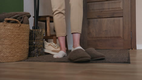 patient stands in cozy room after removing canvas shoes, preparing to wear crocs, wearing beige pants and oversized white sweater, in front of wooden door with green walls, and indoor furniture