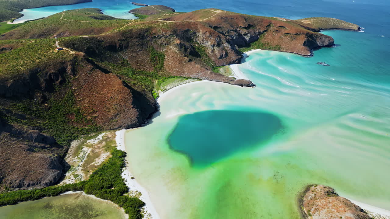 Aerial drone view of a coastal lagoon with turquoise and green tones surrounded by dry rocky hills and vegetation in Baja California Sur