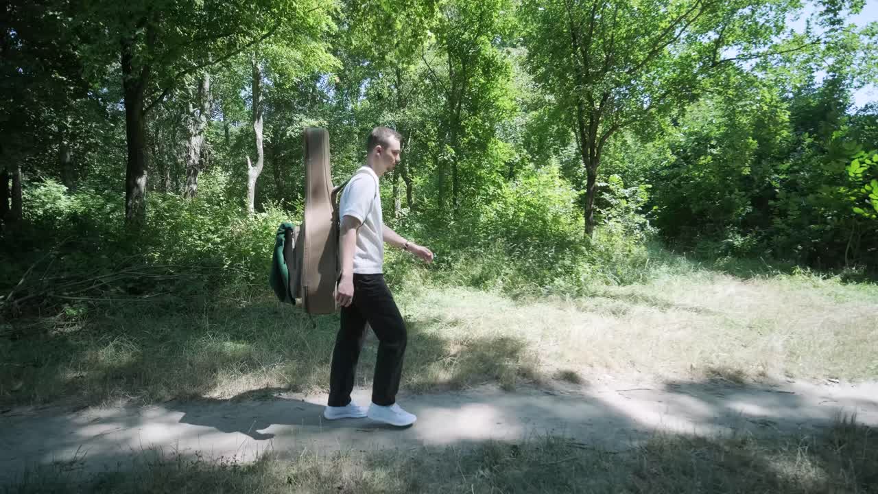 Nuori mies kävelemässä kitaran kanssa kadulla metsän lähellä