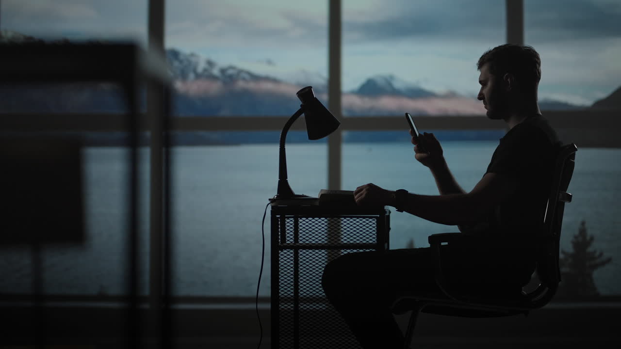 Silhouette of a man sitting at a table looking at a mobile phone thinking and analyzing the stock market solving personal problems repayment of loans Internet banking