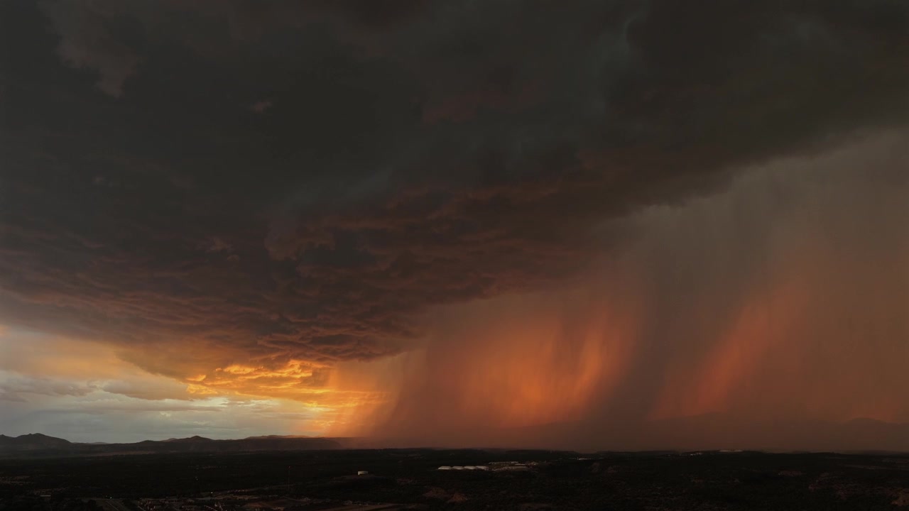 4K aerial shot capturing extreme weather. A massive, heavy monsoon storm drops a torrential downpour over a dark landscape. The thick rain curtain glows fiery orange, backlit by intense light.