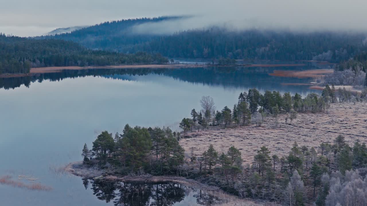 En rolig innsjø reflekterer den tåkete skogen og bølgende åser under en myk grå himmel – luftfoto med drone