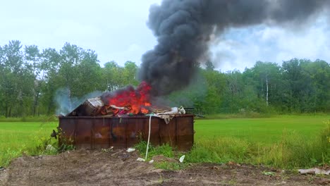 thick dark smoke billowing from a burning dumpster full of garbage on rural farmland