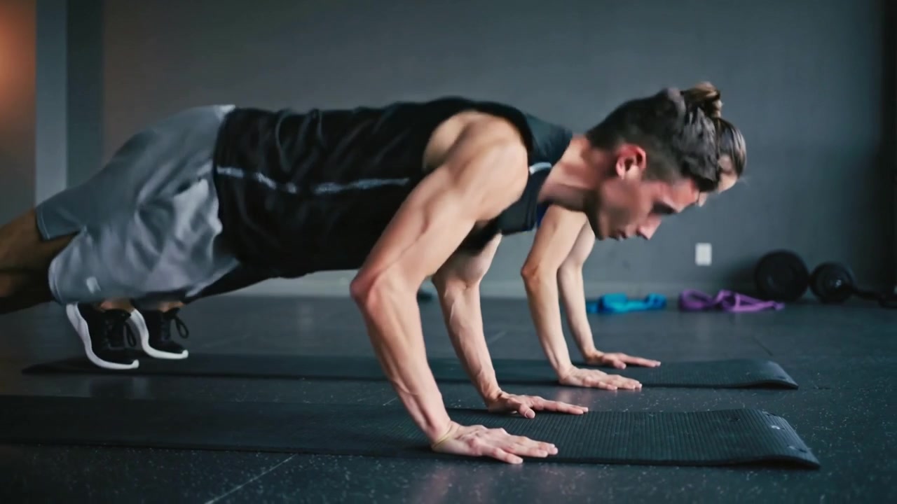 Partner che partono da una posizione bassa, saltando in posizione plank e abbassando il petto su tappetini neri, indossando abiti neri e blu.