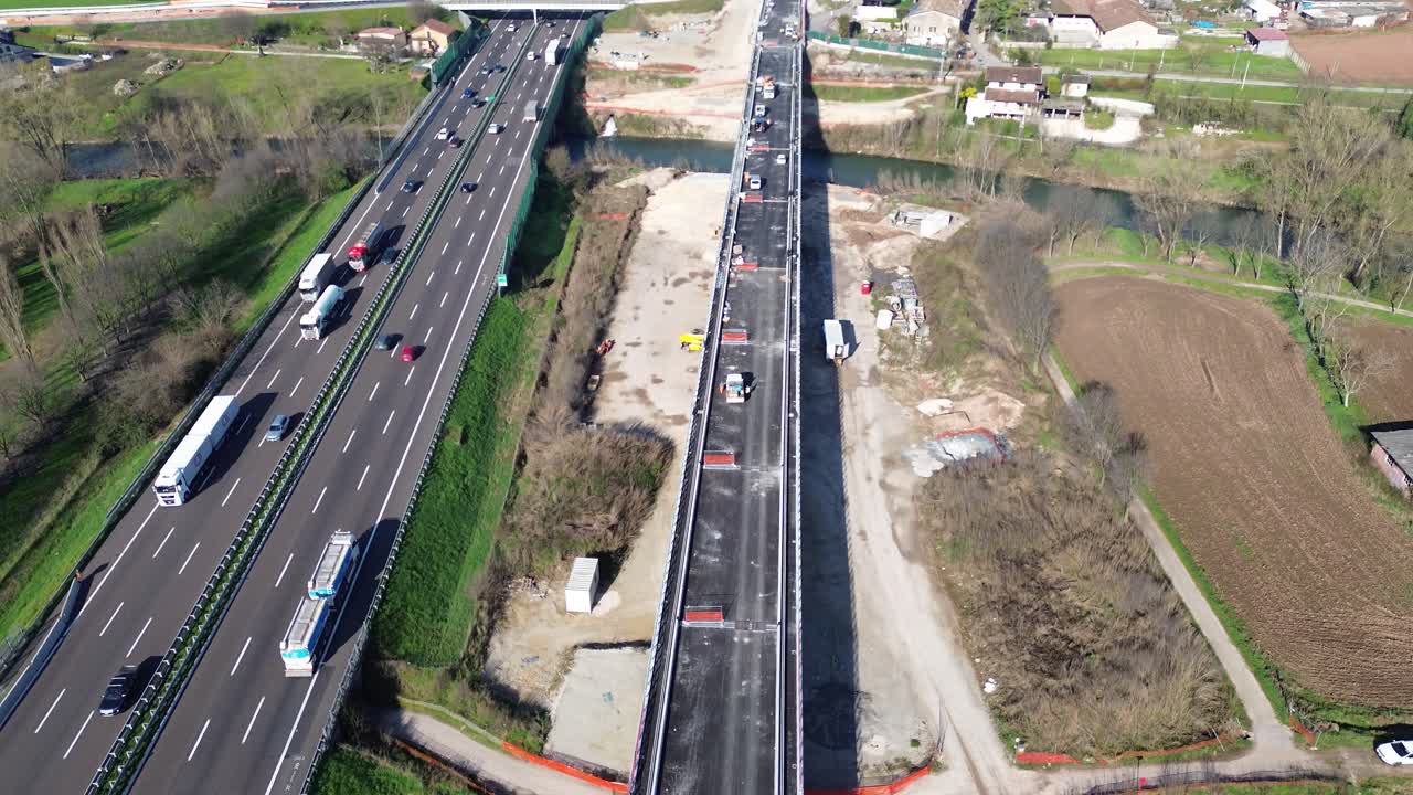 High-resolution aerial footage showing a busy Italian highway with dense traffic alongside an active construction site for a high-speed rail (TAV) project.
