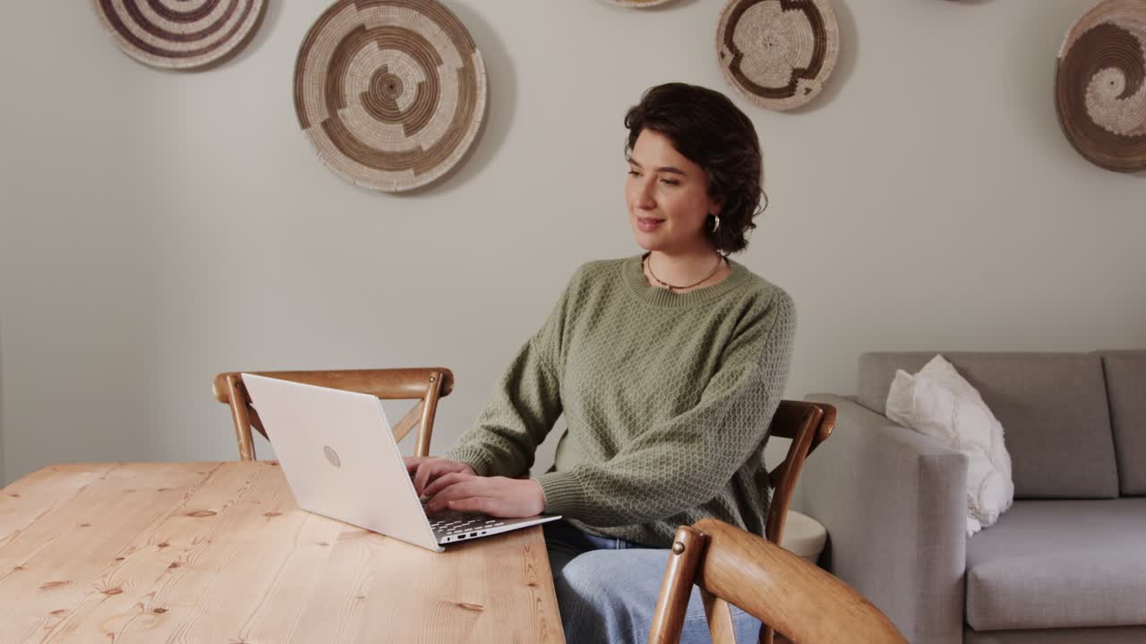 Woman typing on silver laptop in dining area, seeing funny message on screen, laughing while typing