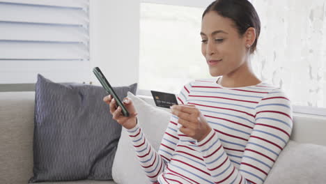 mujer biracial feliz sentada en el sofá usando teléfono inteligente para comprar en línea, cámara lenta