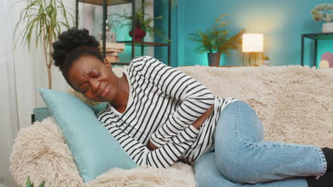 african american woman feeling stomach pain holding abdomen lying exhausted on couch indoors