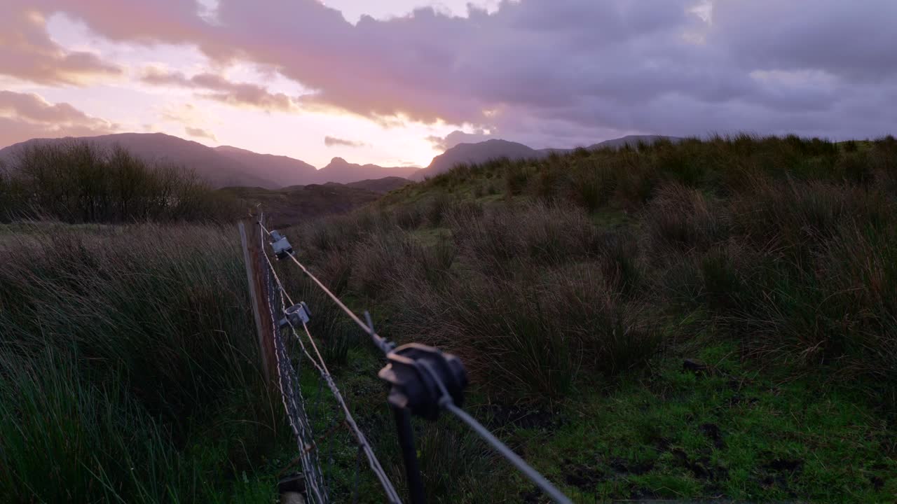 таймлапс розовых и желтых облаков, пролетающих мимо при сильном ветре на закате, с забором для скота и пучками травы на переднем плане и горами в силуэте на заднем плане.