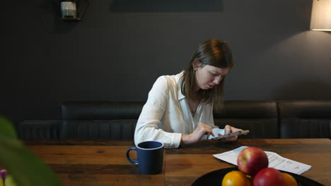 Una-Mujer-Joven-Enviando-Mensajes-De-Texto-A-Su-Teléfono-En-Una-Cocina-Con-Una-Carta-Y-Papeleo-Sobre-La-Mesa