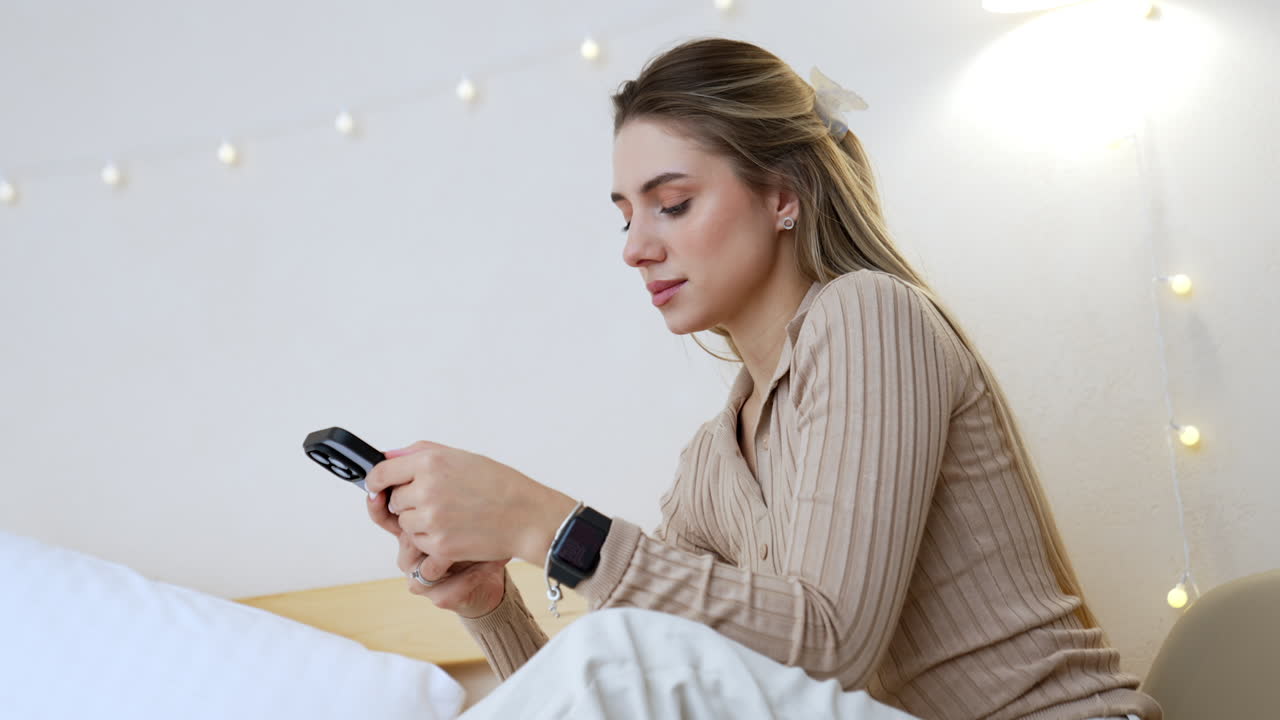 Long-haired Caucasian blonde sits indoors using her phone. Side view portrait of the woman focused on her gadget. Low angle view.