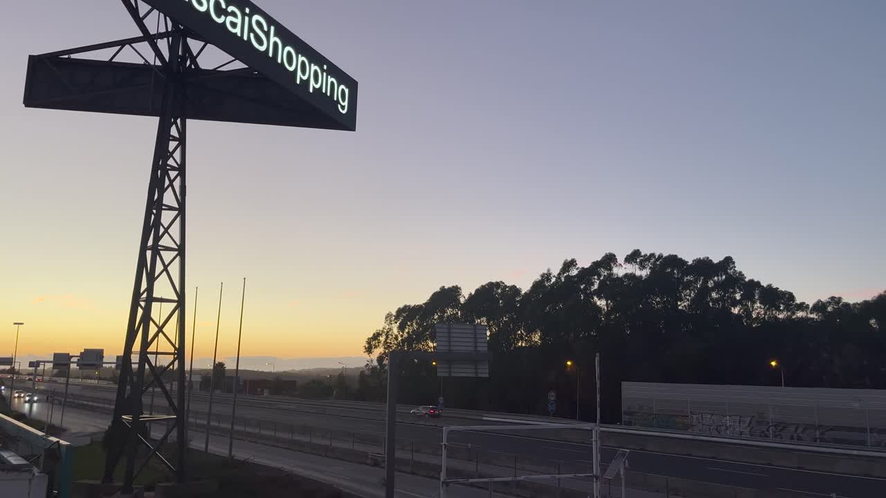 Cielo naranja vibrante al atardecer sobre una autopista concurrida, con una gran valla publicitaria que dice 'Shopping' destacando en la vista panorámica