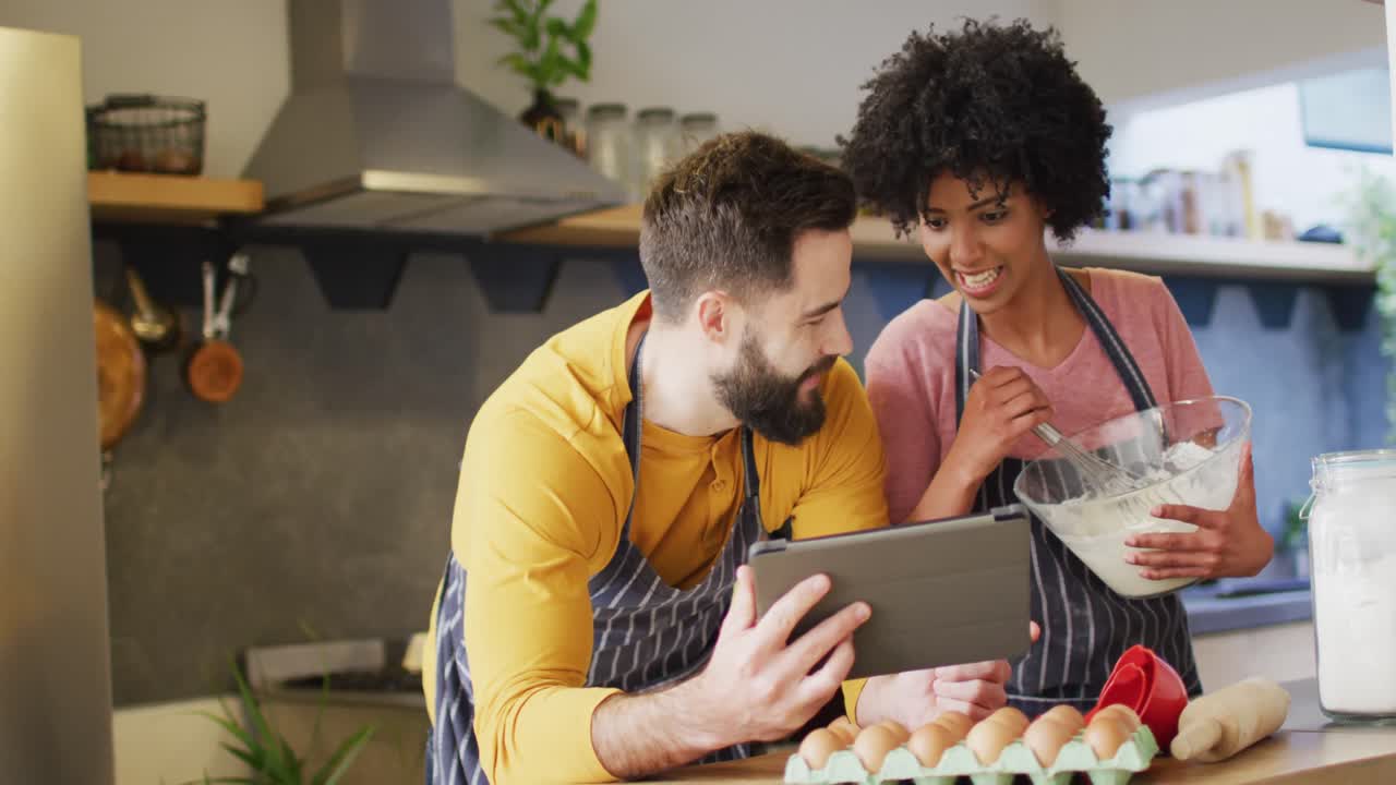 video de una feliz pareja diversa en delantales usando una tableta, horneando juntos en la cocina, con espacio para copiar