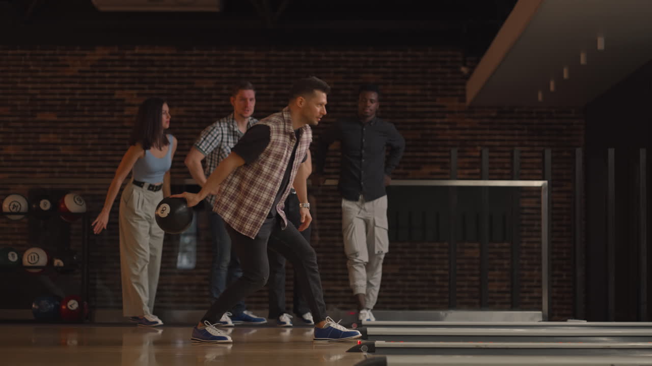 A young Caucasian man makes the final throw with a bowling ball and wins the game with the support and joy of his friends of different nationalities. Rejoice and celebrate the victory
