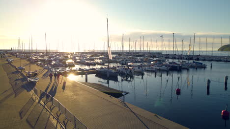 aerial view of people walking along ocean harbor with tall sailboats at sunset