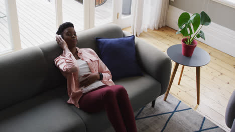mujer afroamericana escuchando música en auriculares mientras se acuesta en el sofá en casa