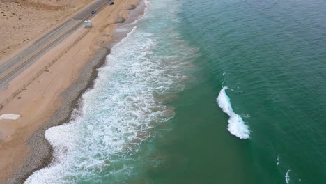 flying along shoreline in malibu, aerial shot ocean waves crashing against sand, cars moving along highway