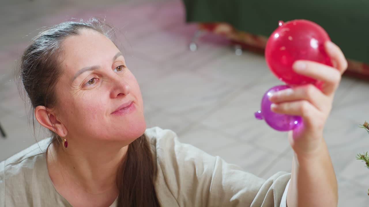 Close up view of woman smiling gently while holding colorful red and purple ornaments in hand, examining holiday decorations closely with admiration, surrounded by cozy festive home atmosphere