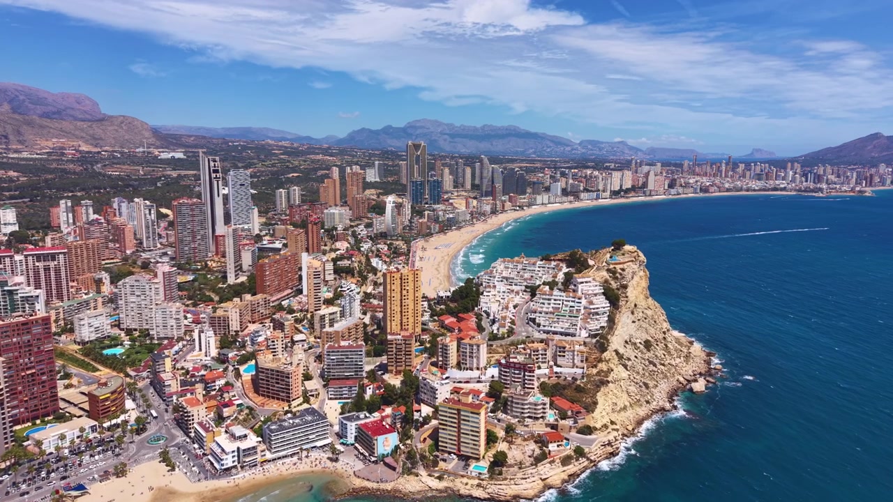Benidorms kystlinje i Spanien, der viser skyskrabere, hoteller, buet sandstrand, turkis Middelhav, klippefyldt forbjerg og bjerge i baggrunden på en solrig sommerdag