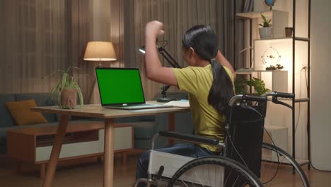 back view of asian kid girl sitting in a wheelchair while using laptop computer with mock up green screen and celebrating at home