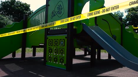 yellow vibrant colored crime scene tape with do not enter written on it moving in front of a childrens playground that has a small slide that is shiny during a warm and sunny summer day. investigation