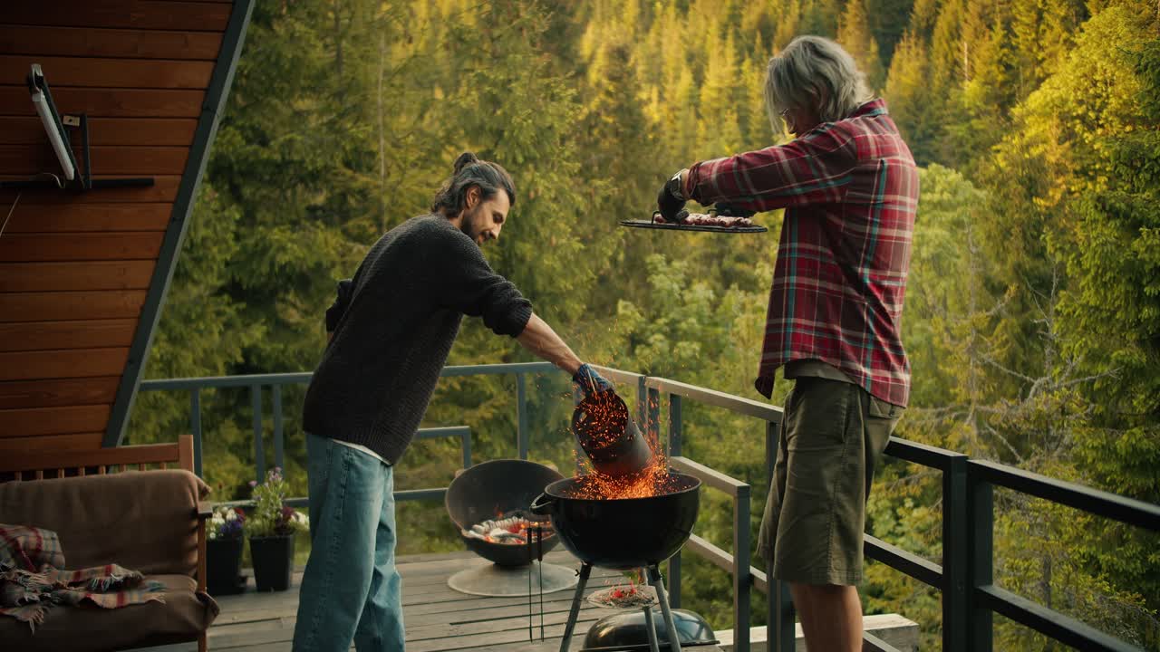 금발 남자와 갈색 남자가 그릴에서 음식을 요리합니다. 빨간색 색 셔츠를 입은 남자는 그릴을 올려서 초록색 스웨트 셔츠에 입은 남자가 뜨거운 석탄을 부어줍니다. 산과 나무 숲의 전망으로 피크닉