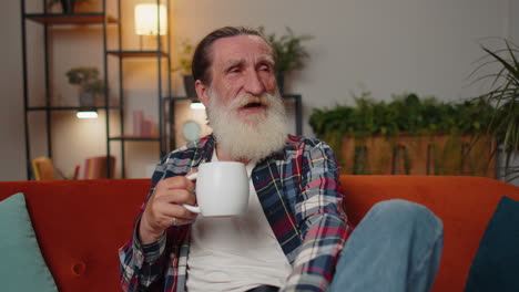 Attractive-smiling-senior-old-grandfather-man-drinking-a-cup-of-coffee-or-herbal-tea-at-home-couch