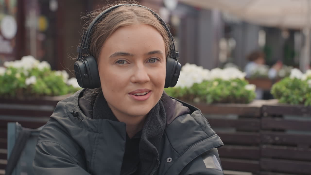 Ung kvinne som lytter til musikk i hodetelefoner på en utendørs kafé, iført svart jakke på en terrasse med potteplanter, ærlige nærbilder viser et avslappet smil og subtil latter, øynene lukket mens hun føler musikken, myk høst