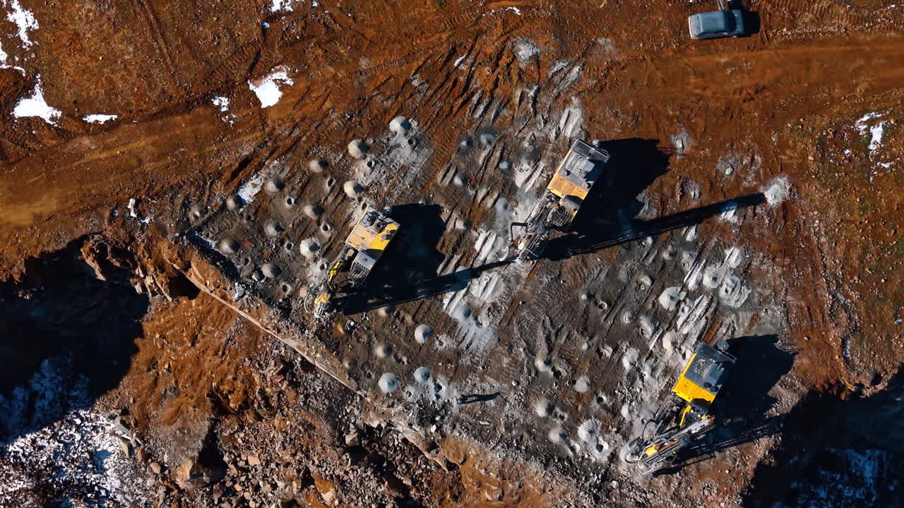 Top down view of drill rigs on quarry bench. Three yellow hydraulic crawler drills boring holes into rock surface for blasting at a granite mine
