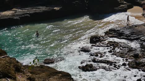 Gente-Nadando-En-Las-Olas-Y-Las-Rocas-Y-De-Pie-En-La-Arena-De-Una-Playa-En-Oahu-Hawaii-Con-Roca-Volcánica-Negra-Por-Todas-Partes