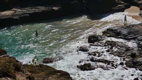 people swimming in the rough surf and rocks and standing on the sand of a beach on oahu hawaii with black volcanic rock all around